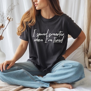 May include: Dark gray t-shirt with the text "I sound smarter when I'm tired" in white script. The shirt is worn with blue jeans. A white vase with cotton stems is in the background.