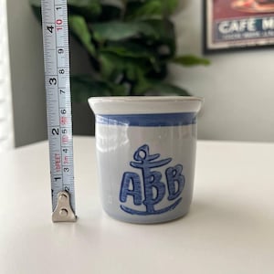 May include: A light gray ceramic mug with a blue rim and a blue anchor graphic featuring the letters ABB. A metal measuring tape is positioned vertically next to the mug, indicating a height of approximately 2.75 inches for the ceramic vessel.