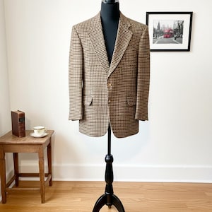 May include: A brown and tan houndstooth blazer displayed on a black mannequin. The blazer has two front pockets and two buttons. The mannequin is on a black stand. A small wooden table with a book and teacup is to the left.