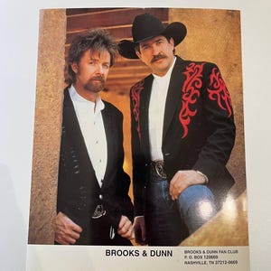 May include: A promotional photograph of the country music duo Brooks & Dunn. One individual wears a black blazer, while the other wears a black blazer with red flame-like embroidery. Both wear white shirts. Text at the bottom reads BROOKS & DUNN.