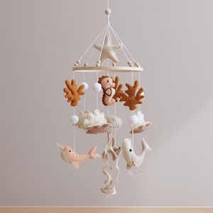 May include: A hanging mobile featuring felt sea creatures and ocean elements suspended from a circular wooden frame. The mobile includes a beige starfish, a seahorse, coral shapes in rust orange, white felt balls, seashells, a narwhal, and a whale.