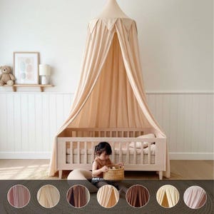 Puede incluir: Una cama con dosel beige con estructura de madera. El dosel está drapeado sobre la cama, creando un espacio acogedor. Un niño está sentado cerca de la cama, sosteniendo una cesta. La habitación tiene una paleta de colores neutros.