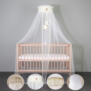 Puede incluir: Una cuna de madera clara con un dosel de mosquitera blanco. El dosel tiene una parte superior de color crema con un diseño de oso y cintas colgantes con adornos de estrella y luna. La cuna está vacía, con un colchón blanco.