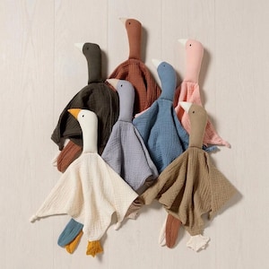 May include: A collection of seven soft goose-shaped security blankets made from textured muslin fabric. Each plush toy features a long neck, a small head with a simple eye, and a draped body in various colours including white, olive, rust, blue, and pink.