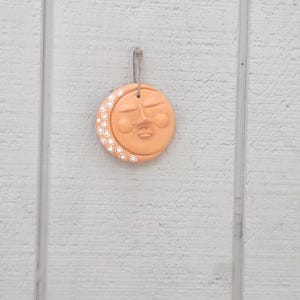 May include: A circular peach-colored clay ornament featuring a sculpted face with closed eyes and a small mouth. The left side of the crescent moon shape is decorated with a pattern of small white dots. A thin twine loop is attached for hanging.