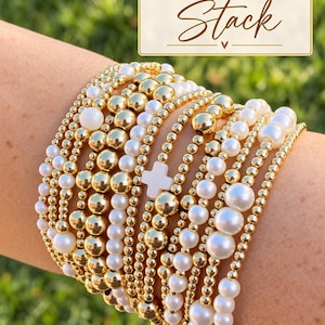 May include: A stack of multiple beaded bracelets featuring alternating gold-tone metallic spheres and white spherical beads of varying sizes. One bracelet includes a white cross-shaped charm. The image includes the text Build Your Own Stack.