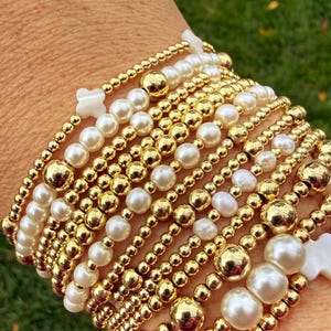 May include: A stack of multiple stretch bracelets featuring a combination of polished gold-tone metal beads, white round pearls, and small white cross-shaped beads. The bracelets vary in bead size and arrangement, creating a layered appearance.