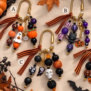 May include: Four gold-toned metal carabiner keychains labeled A, B, C, and D, each featuring orange and black striped ribbons and assorted Halloween-themed beads and charms, including pumpkins, ghosts, bats, skulls, and skeletons.