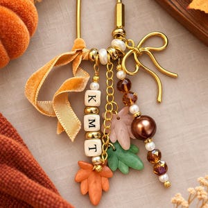 May include: A gold-toned metal keychain featuring a U-shaped clasp, a bow charm, and multiple dangling elements including white square beads with the letters K, M, and T, orange and green leaf-shaped charms, brown and white beads, and a velvet ribbon.