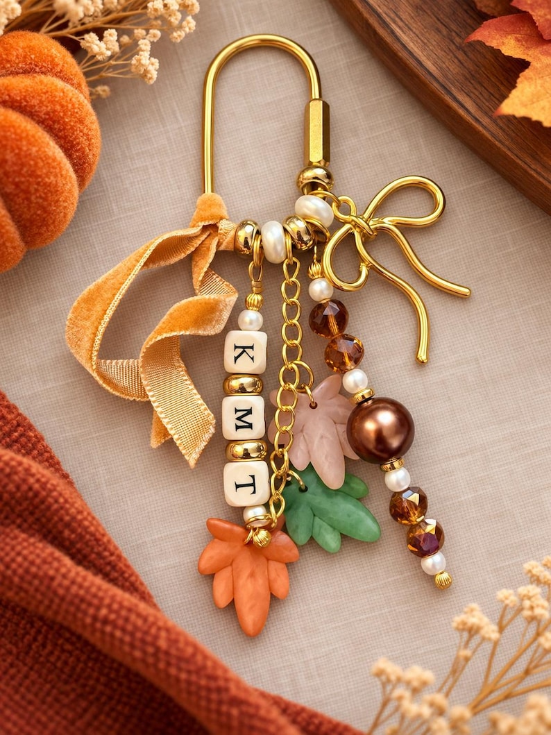 May include: A gold-toned metal keychain featuring a U-shaped clasp, a bow charm, and multiple dangling elements including white square beads with the letters K, M, and T, orange and green leaf-shaped charms, brown and white beads, and a velvet ribbon.