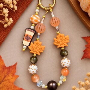 May include: A beaded keychain featuring a gold-toned ring, orange and white checkered beads, translucent orange striped beads, a coffee cup charm with a maple leaf graphic, orange maple leaf beads, and various brown, white, and floral beads.