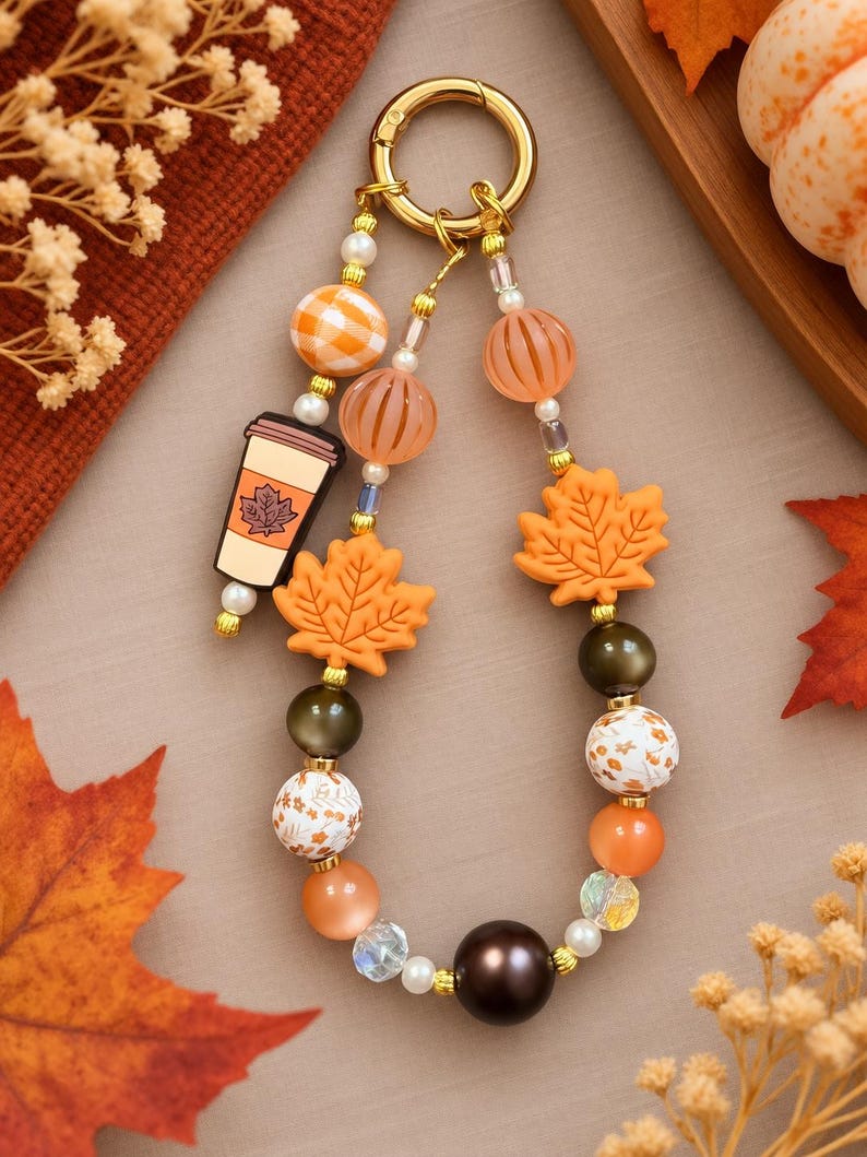May include: A beaded keychain featuring a gold-toned ring, orange and white checkered beads, translucent orange striped beads, a coffee cup charm with a maple leaf graphic, orange maple leaf beads, and various brown, white, and floral beads.