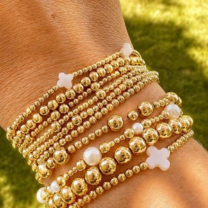 May include: A stack of multiple stretch bracelets featuring polished gold-tone metallic beads of varying sizes, interspersed with smooth white spherical pearls and small white four-petaled flower-shaped charms arranged in a layered design.