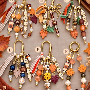 May include: Six gold-toned metal keychains labeled A through F, featuring assorted beads in orange, brown, and cream, fabric ribbons, and charms including maple leaves, mushrooms, and coffee cups. Some keychains include lettered wooden beads.