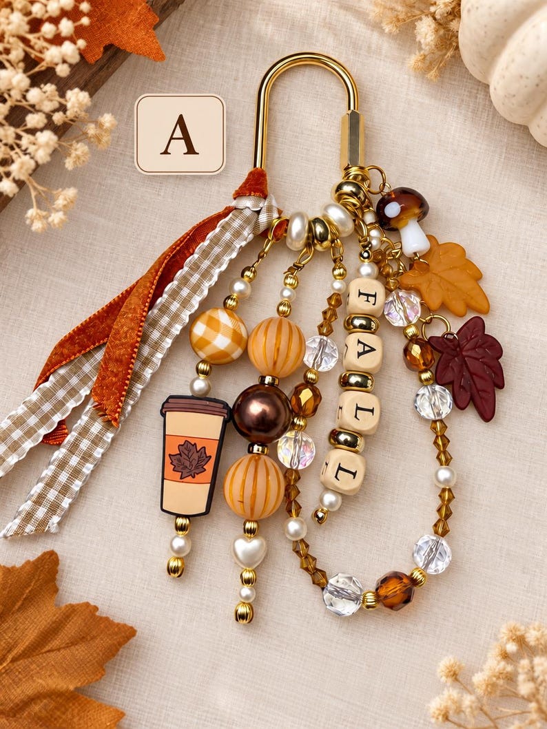 May include: A gold-toned metal clasp keychain featuring a collection of charms including a coffee cup, a mushroom, and maple leaves. The keychain includes wooden beads spelling FALL, orange and brown striped beads, and ribbons in plaid.