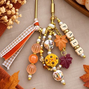 May include: A gold-toned metal carabiner keychain featuring a dangling assortment of beads, including orange striped spheres, a yellow sunflower charm, and white beads with printed imagery. A strand of wooden letter beads spells out FALL.