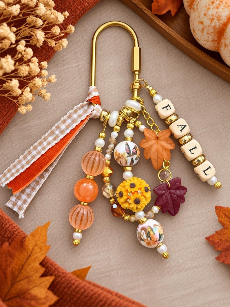 May include: A gold-toned metal carabiner keychain featuring a dangling assortment of beads, including orange striped spheres, a yellow sunflower charm, and white beads with printed imagery. A strand of wooden letter beads spells out FALL.