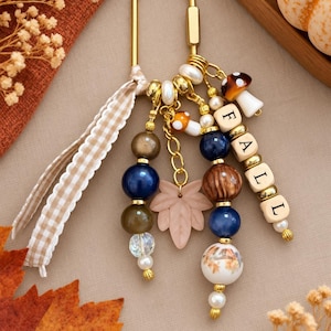 May include: A gold-toned metal keychain featuring a variety of beads in shades of blue, brown, and white, along with a light pink leaf charm, two small mushroom charms, and square letter beads spelling the word FALL, accented by a gingham ribbon.