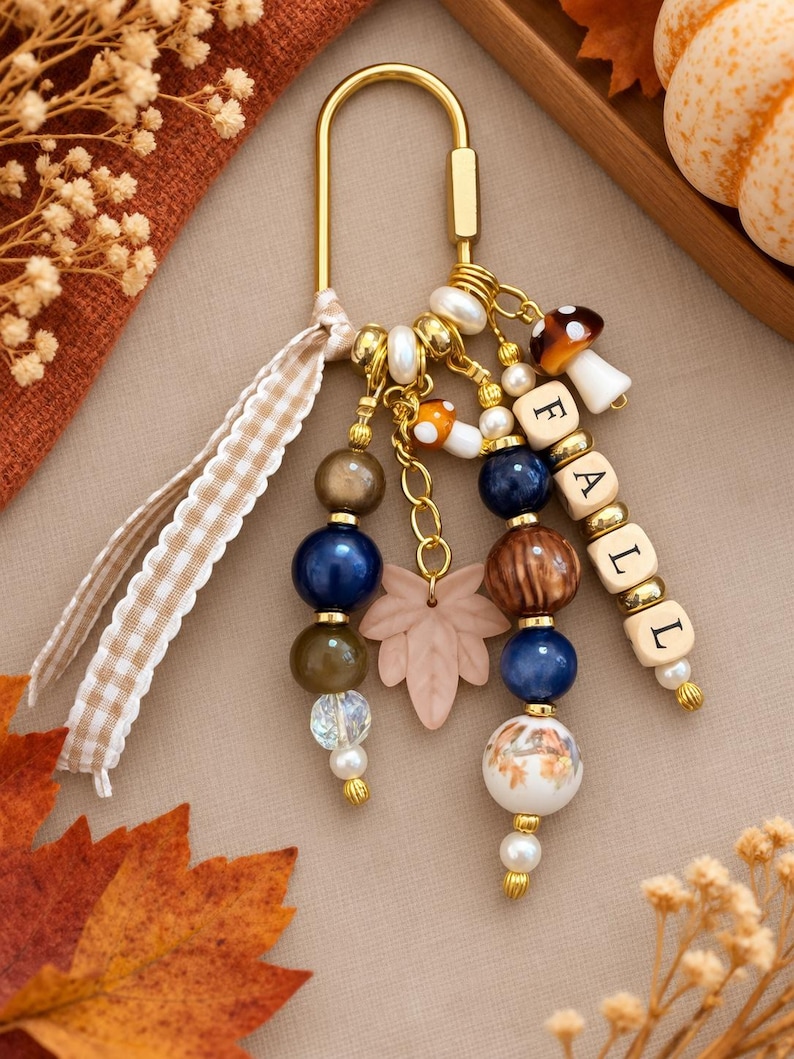 May include: A gold-toned metal keychain featuring a variety of beads in shades of blue, brown, and white, along with a light pink leaf charm, two small mushroom charms, and square letter beads spelling the word FALL, accented by a gingham ribbon.