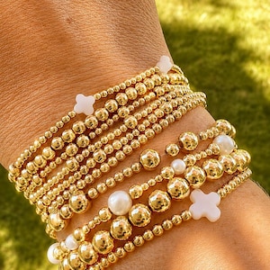 May include: A stack of multiple stretch bracelets featuring polished gold-tone metallic beads of varying sizes, interspersed with smooth white spherical beads and small white four-petaled flower-shaped charms arranged in a layered design.