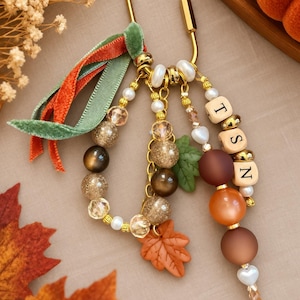 May include: A gold-toned metal carabiner keychain featuring a collection of dangling beads in shades of brown, orange, and cream. The accessory includes a green velvet ribbon, a maple leaf charm, and three square beads displaying the letters T, S, and N.
