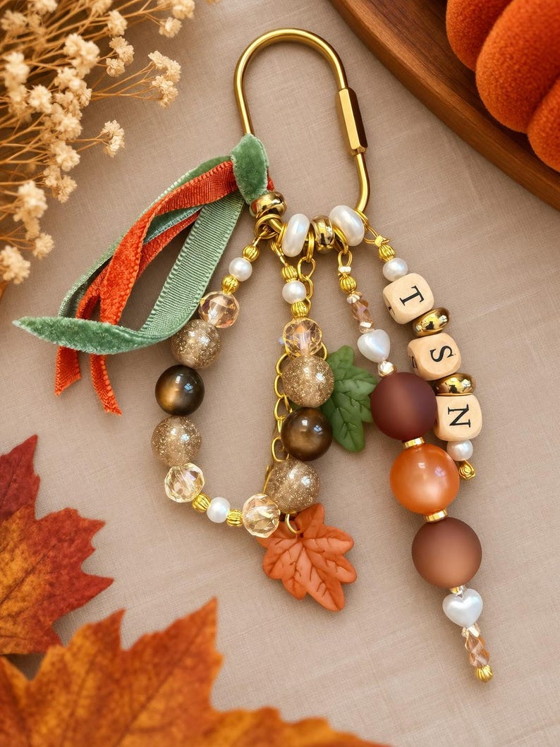 May include: A gold-toned metal carabiner keychain featuring a collection of dangling beads in shades of brown, orange, and cream. The accessory includes a green velvet ribbon, a maple leaf charm, and three square beads displaying the letters T, S, and N.