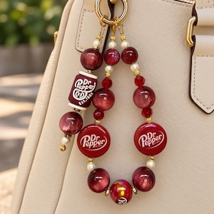 May include: A decorative bag charm featuring maroon and white beads, including a cylindrical bead with Dr. Pepper text and two circular Dr. Pepper logo charms. The charm is attached to a gold-toned metal ring and hangs from a cream-colored handbag.
