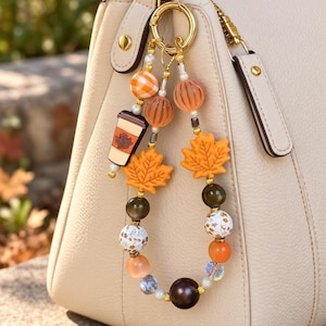 May include: A decorative bag charm featuring a gold-toned ring with dangling beads in shades of orange, brown, and white. The charm includes silicone maple leaf shapes, a coffee cup pendant, and various round beads with plaid and floral patterns.