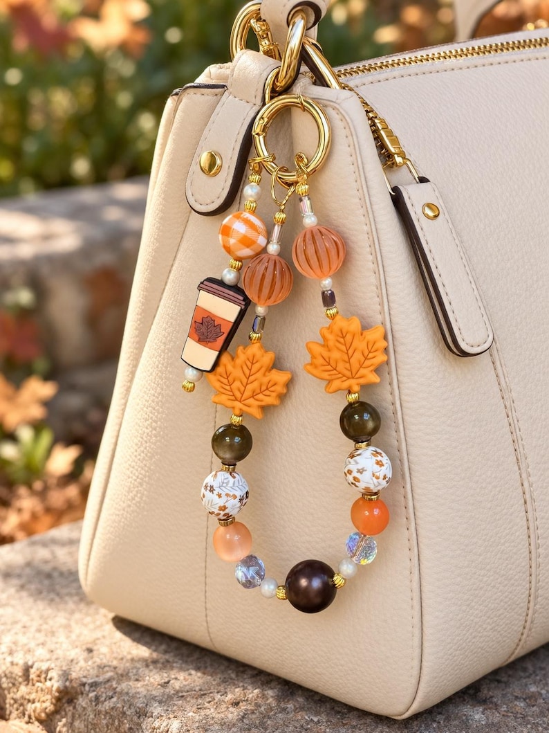 May include: A decorative bag charm featuring a gold-toned ring with dangling beads in shades of orange, brown, and white. The charm includes silicone maple leaf shapes, a coffee cup pendant, and various round beads with plaid and floral patterns.