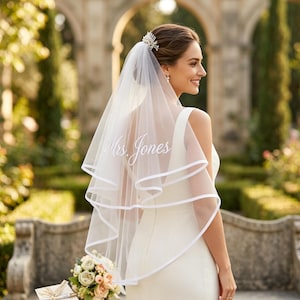 Pode incluir: Uma noiva usando um véu de noiva branco com o nome "Mrs. Jones" bordado. O véu tem um design de camada dupla com acabamento branco. A noiva está usando um vestido branco e um acessório de cabelo. Um buquê de flores e um cartão estão no fundo.