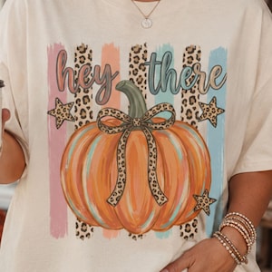 Peut inclure: Un t-shirt à manches courtes de couleur crème arborant un motif de citrouille orange avec un nœud imprimé léopard, entouré de rayures verticales pastel, d'accents léopard et d'étoiles, avec le texte "hey there" imprimé au-dessus.