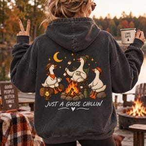 Peut inclure: Un sweat à capuche gris anthracite avec un motif au dos représentant trois oies blanches assises autour d'un feu de camp. Les oies portent des chapeaux et tiennent des tasses et des guimauves. Le texte indique "JUST A GOOSE CHILLIN".