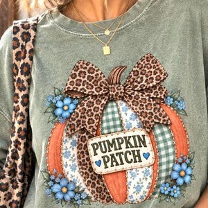 Peut inclure: Un sweat-shirt col rond vert sauge avec un motif central de citrouille composé de panneaux orange, à fleurs bleues et à carreaux verts. La citrouille est surmontée d'un nœud léopard et de fleurs bleues, avec l'inscription PUMPKIN PATCH.