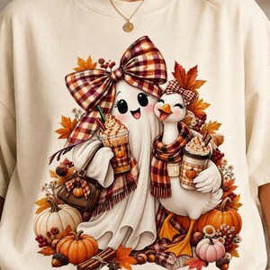 A person wearing a beige t-shirt featuring a ghost and a duck with coffee cups, plaid bows, and pumpkins.