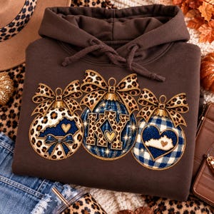 May include: A dark brown hooded sweatshirt featuring a graphic of three Christmas ornaments. The ornaments are decorated with leopard print, blue plaid, and glittery textures, displaying the letters KY and the outline of the state of Kentucky.