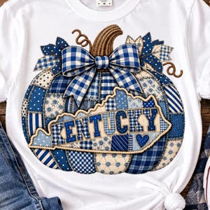 May include: A white t-shirt featuring a large graphic of a pumpkin made from various blue and white patterned fabric patches. The pumpkin is topped with a blue and white gingham bow and includes a map outline of Kentucky with the text KENTUCKY.