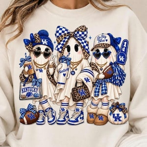 May include: A cream-colored sweatshirt featuring a graphic of three ghosts dressed in University of Kentucky apparel. The ghosts wear blue and leopard print accessories, hold items like a football and a cup, and display the text Kentucky and Go Big Blue.