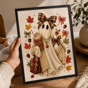 A person holding a framed print displaying an illustration of a ghost in a plaid bow and sweater, a goose, a latte, autumn leaves, and acorns against a white background.