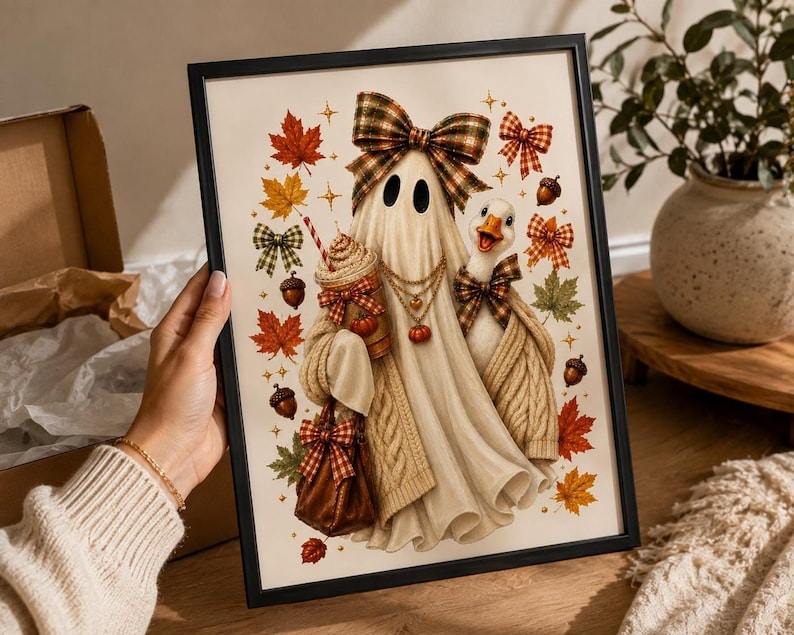 A person holding a framed print displaying an illustration of a ghost in a plaid bow and sweater, a goose, a latte, autumn leaves, and acorns against a white background.
