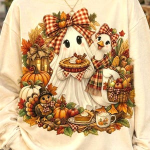 A person wearing a beige sweatshirt displaying a ghost holding a pie and a duck next to a Thanksgiving-themed harvest wreath.