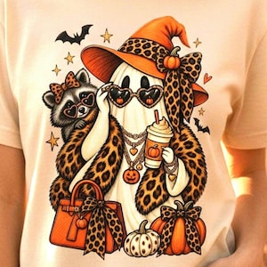 Peut inclure: Un t-shirt couleur crème avec un motif représentant un fantôme portant un chapeau à imprimé léopard, des lunettes de soleil en forme de cœur et un châle léopard. Le fantôme tient un café et un raton laveur, entouré de citrouilles et d'étoiles.