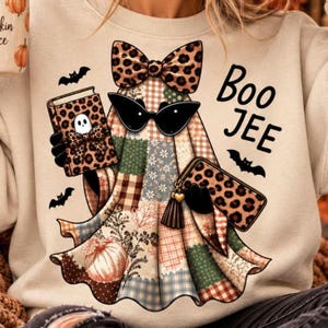 Peut inclure: Un sweat-shirt beige avec un motif de fantôme portant des lunettes de soleil œil-de-chat noires et un nœud à imprimé léopard. Le fantôme tient un livre et un portefeuille léopard, avec des citrouilles, des chauves-souris et le texte Boo JEE.