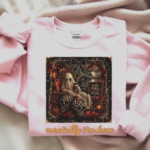 Peut inclure: Un sweat-shirt à col rond rose clair avec une impression graphique représentant un fantôme assis sur une chaise avec des crânes, une lanterne et un panneau indiquant hello FALL COZY DAYS WARM DRINKS & GOOD VIBES. Le texte mentally i'm here est imprimé en dessous.