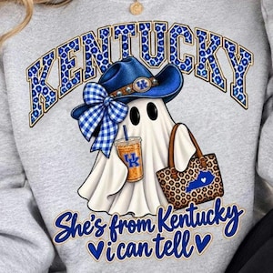 Peut inclure: Un sweat-shirt gris avec un motif de fantôme blanc portant un chapeau de cow-boy bleu avec un nœud à carreaux bleu et blanc. Le fantôme tient un gobelet à café et un sac fourre-tout à motif léopard. Le texte indique KENTUCKY et She's from Kentucky I can tell.