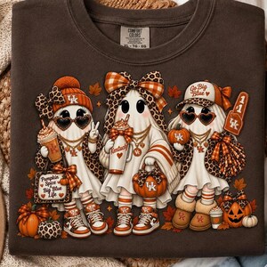 Può includere: Una felpa girocollo marrone scuro con una grafica di tre fantasmi che indossano accessori autunnali, tra cui fiocchi leopardati, berretti arancioni e cappellini. I fantasmi tengono in mano tazze di caffè e zucche con il marchio Kentucky e foglie autunnali.