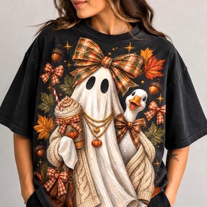 Peut inclure: Un t-shirt noir à manches courtes orné d'un motif représentant un fantôme portant un gilet en tricot et des chaînes dorées, tenant un café. À côté du fantôme se trouve une oie avec un nœud à carreaux, des feuilles d'automne et des glands.