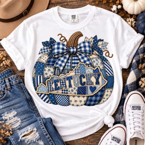 Peut inclure: Un t-shirt blanc à manches courtes orné d'un grand motif de citrouille composé de pièces en patchwork bleu et blanc. Un nœud en vichy bleu et blanc surmonte la citrouille, qui présente le contour de l'État du Kentucky avec le texte KENTUCKY.