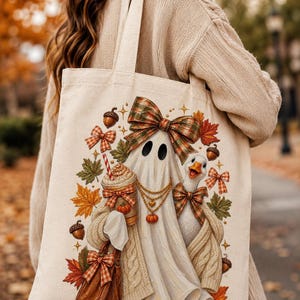 A person wearing a cream sweater carrying a light beige tote bag printed with an illustration of a ghost, a goose, autumn leaves, and acorns.