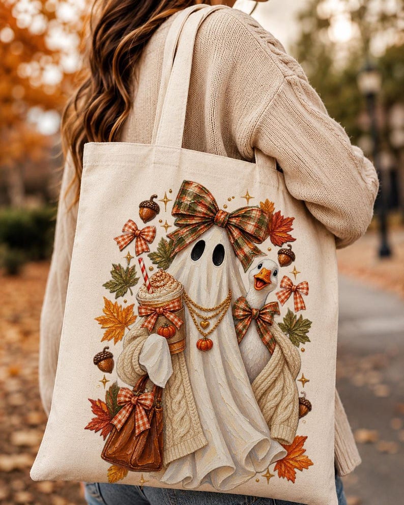 A person wearing a cream sweater carrying a light beige tote bag printed with an illustration of a ghost, a goose, autumn leaves, and acorns.