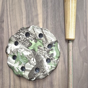May include: A wooden duck call with a camouflage base and a light-colored wooden handle. The call has a multi-colored striped stem. The camouflage base has several black holes.